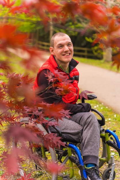 Portrait of a paralyzed young man in a red vest in a public park in the city Sitting in the wheelchair next to autumn leaves