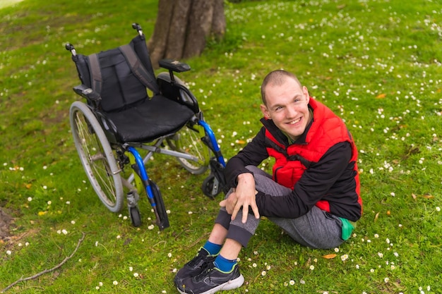 Portrait of a paralyzed young man sitting on the grass next to the wheelchair in the spring in a natural park