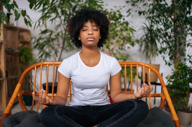 Portrait of peaceful black young woman sitting with closed eyes on sofa in lotus pose practicing yoga meditation at home relaxing and breathing deep