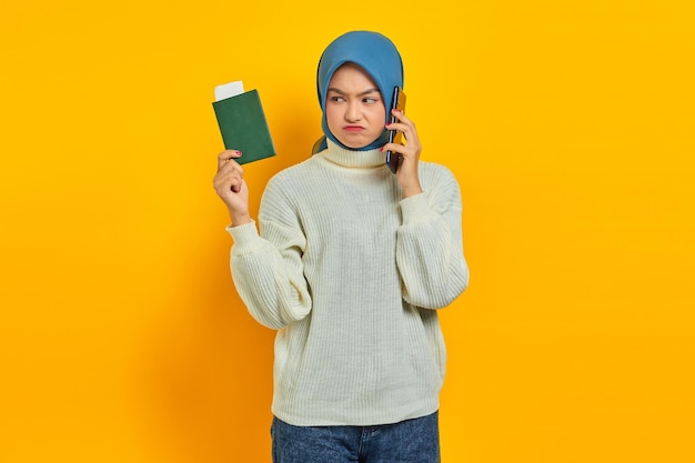 Portrait of pensive beautiful Asian woman in white sweater talking on mobile phone and holding passport isolated over yellow background Summer Trip