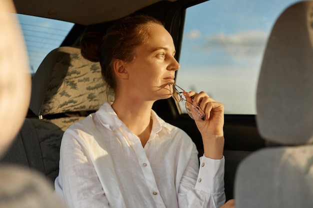 Portrait of pensive calm relaxed attractive woman sitting on back seat in the car with glasses in hands looking away at window enjoying sunset wearing white shirt