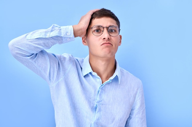 Portrait of pensive man in glasses and shirt touches his head and thinks makes a decision with doubting face isolated in blue studio with copy space