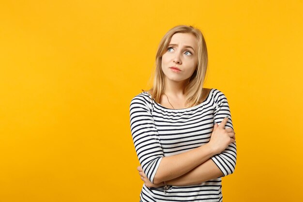 Portrait of pensive young woman in striped clothes looking aside up holding hands crossed isolated on yellow orange background in studio. People sincere emotions lifestyle concept. Mock up copy space.