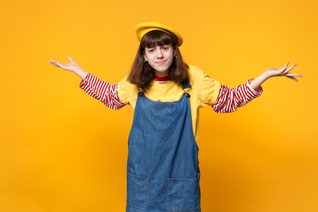 Portrait of perplexed discouraged girl teenager in french beret, denim sundress spreading hands isolated on yellow background in studio. People sincere emotions, lifestyle concept. Mock up copy space.