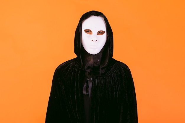 Photo portrait of a person in white killer halloween mask, cape and hood looking at camera, dressed for halloween over orange background