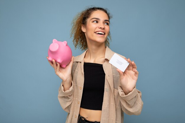 Portrait photo of happy positive smiling young beautiful attractive brunette curly woman with sincere emotions wearing stylish beige shirt isolated over blue background with copy space, holds pink pig