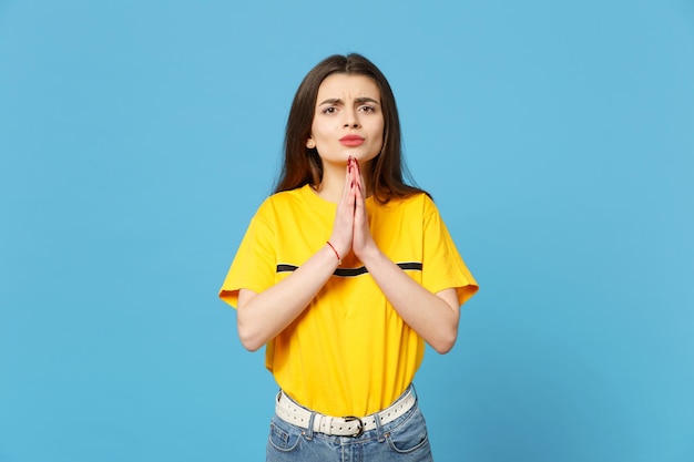 Portrait of pleading young woman in vivid casual clothes looking camera, holding hands folded, praying isolated on bright blue wall background in studio. People lifestyle concept. Mock up copy space.