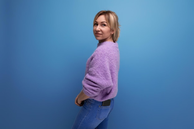 Portrait of a pleasant charming pleasant blond young woman in a purple sweater smiling on a blue