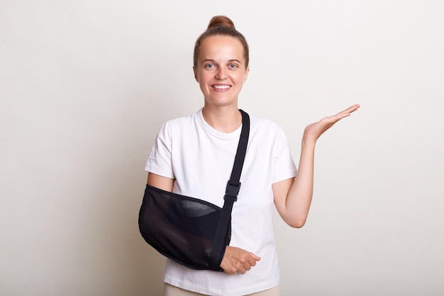 Portrait of pleased adorable woman with bun hairstyle keeps hand spread has smiling expression dressed white Tshirt recovers after accident has broken arm wears sling after visiting surgeon