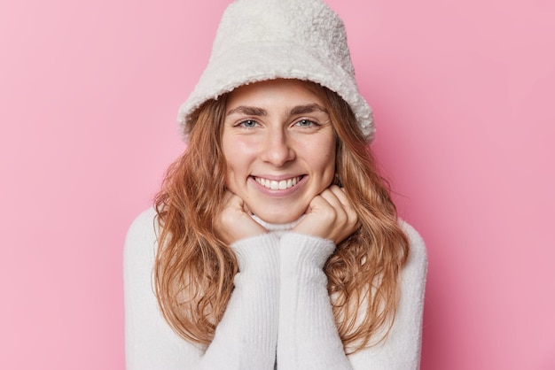 Portrait of pleased beautiful young woman smiles gently keeps hands under chin listens something with pleasure dressed in jumper and winter panama poses against pink background. Happy feelings