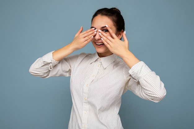 Portrait of positive adult nice winsome brunette woman with sincere emotions wearing white shirt isolated over blue background with copy space and covering eyes with hands