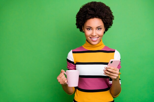 Portrait of positive afro american girl blogger enjoy fall holidays use cellphone follow share repost comment hold hot beverage mug wear bright shine outfit 