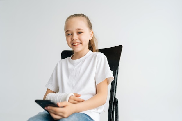 Portrait of positive blonde little girl with broken arm wrapped in white plaster bandage smiling looking at camera holding mobile phone sitting at chair