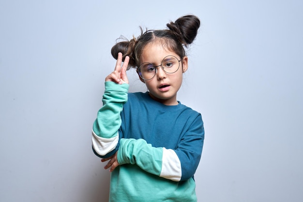 Portrait of positive brunette little girl showing peace gesture in round glasses isolated on white background