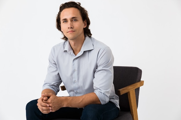 Portrait of positive businessman in office shirt looking at camera while sitting on armchair isolated over white wall