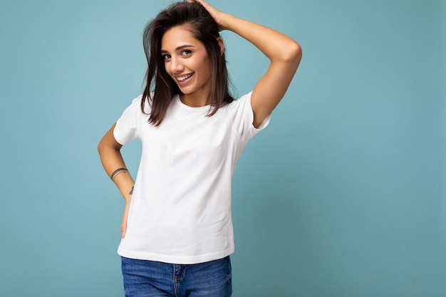 Portrait of positive cheerful fashionable smiling young brunette woman in casual white t-shirt for mockup isolated on blue background with copy space.