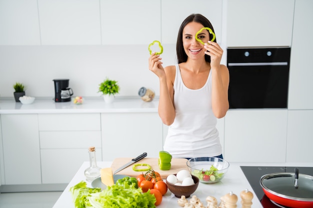 Portrait of positive cheerful girl housewife want be chef prepare vegetarian lunch hold pepper slices imagine it is her glasses wear white singlet in kitchen house indoors