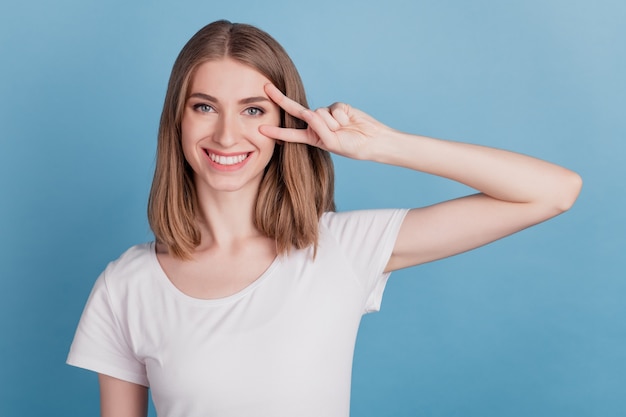 Portrait of positive friendly lady show v-sign cover eye look camera on blue background