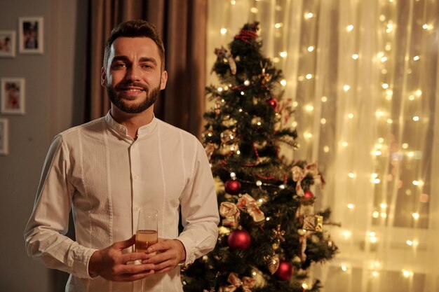 Portrait of positive handsome young man with beard holding champagne flute against Christmas tree in living room