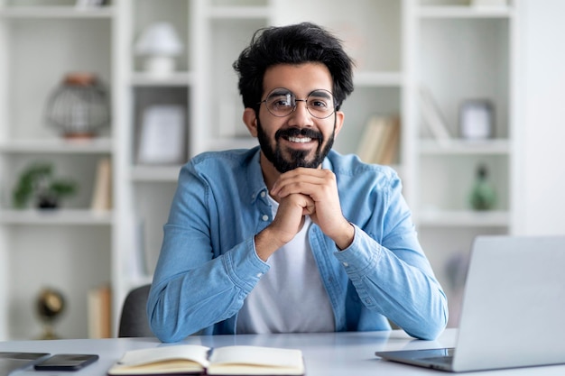 Portrait of positive indian freelancer man sitting at desk in home office