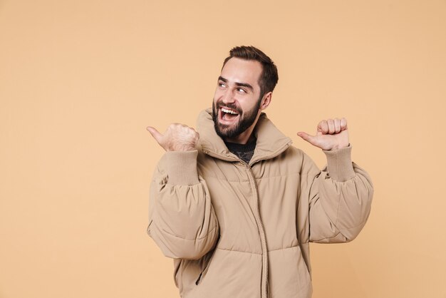 Photo portrait of positive man in winter jacket smiling and pointing fingers aside at copyspace isolated on beige