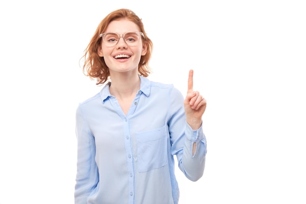 Portrait of a positive redhead young woman in a business shirt smiling pointing her finger at the empty copy space for text or product isolated on white background advertising banner