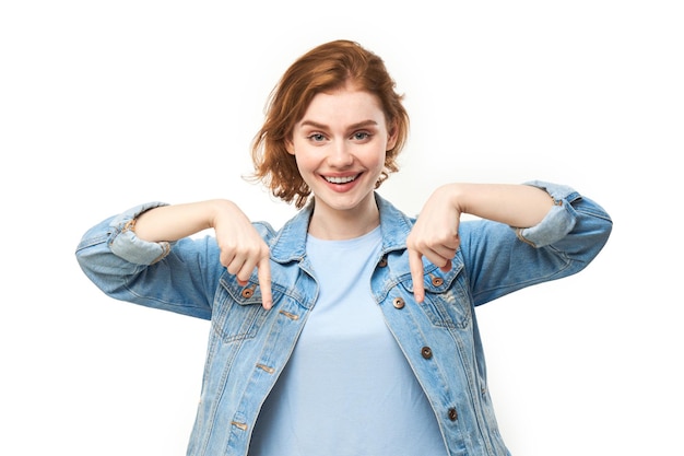 Portrait of positive redhead young woman in casual jeans smiling pointing with finger at empty copy space for text or product isolated on white background advertising banner