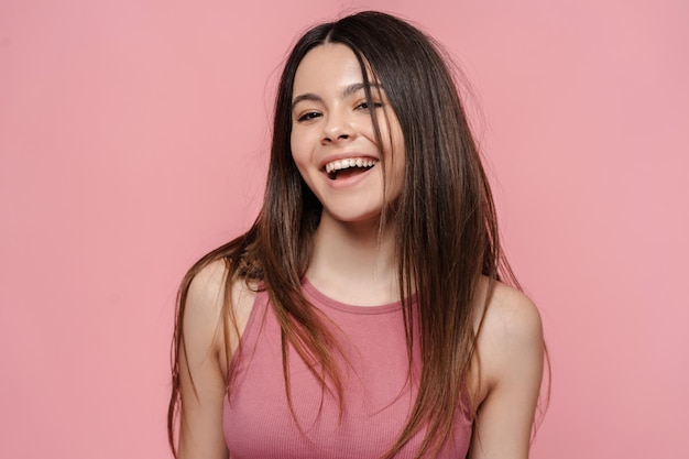 Portrait of positive smiling teenage girl with long hair standing isolated on pink background