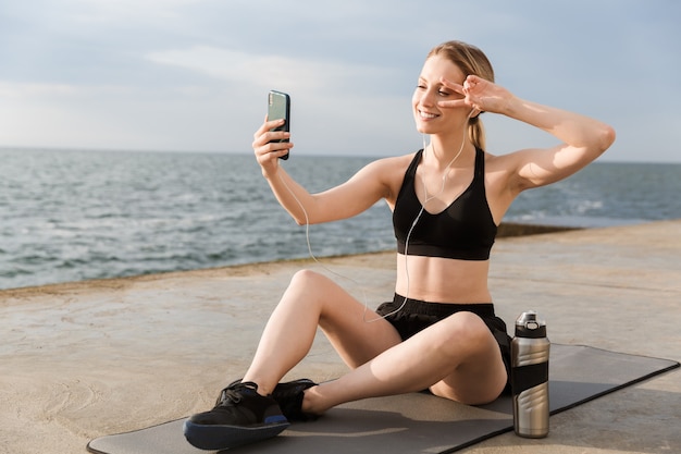 Portrait of positive woman taking selfie on cellphone and gesturing peace sign while sitting on mat near seaside in morning