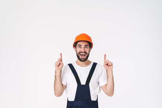 Portrait of positive young man builder in helmet posing isolated over white wall pointing aside.