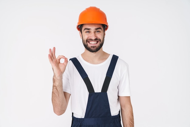 Portrait of a positive young man builder in helmet posing isolated over white wall showing okay gesture.