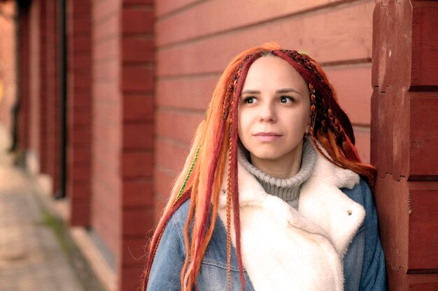 Portrait of positive young woman with dreadlocks standing leaning on wall on city street Pretty female with bright hairstyle looking away smiling