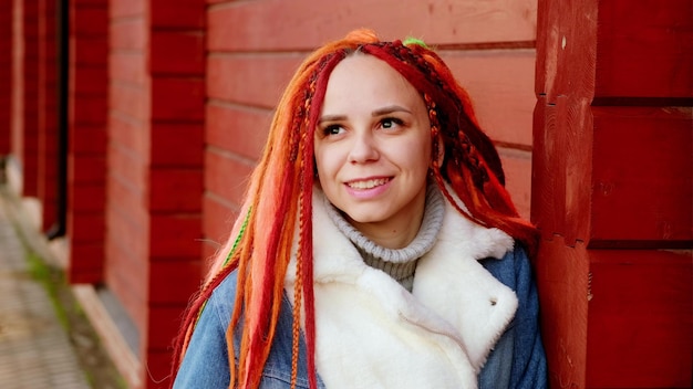 Portrait of positive young woman with dreadlocks standing leaning on wall on city street Pretty female with bright hairstyle looking away smiling