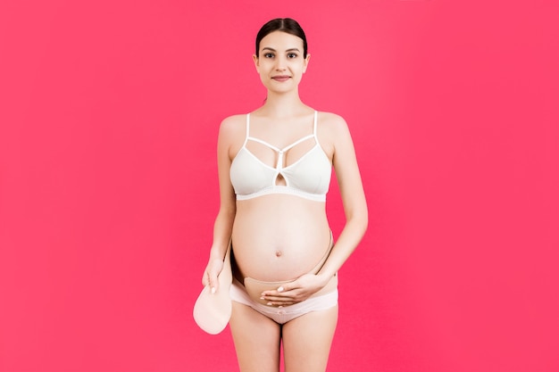 Portrait of pregnant woman in colorful underwear putting on a bandage at pink background with copy space. Orthopedic abdominal support belt concept.