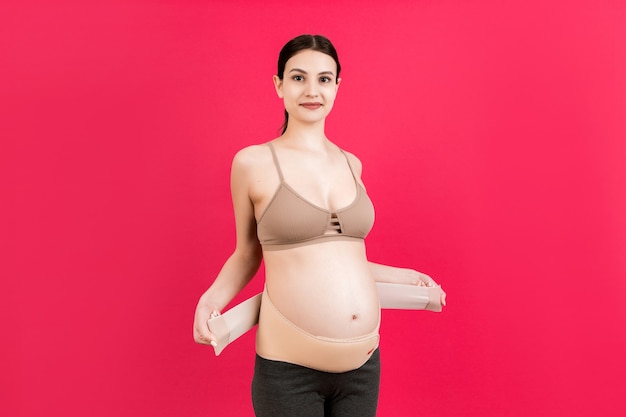 Portrait of pregnant woman putting on supporting bandage for reducing backache at pink background with copy space. Orthopedic abdominal support belt concept.