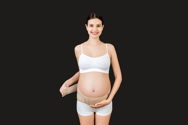 Portrait of pregnant woman in underwear dressing pregnancy corset on the third trimester at black background with copy space. Orthopedic abdominal support belt concept.