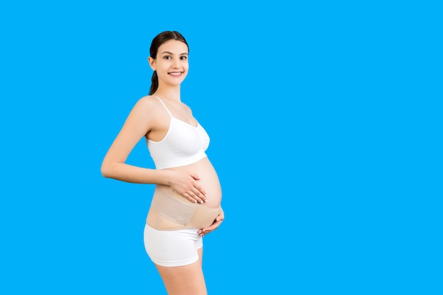 Portrait of pregnant woman in underwear using pregnancy belt at blue background with copy space. Orthopedic abdominal support belt concept.