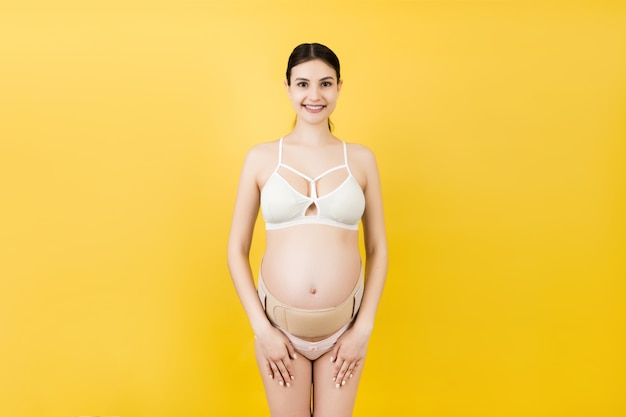 Portrait of pregnant woman in underwear wearing elastic maternity band at yellow background with copy space. Orthopedic abdominal support belt concept.