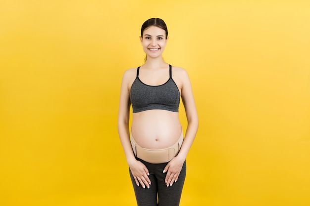 Portrait of pregnant woman wearing pregnancy belt at yellow background with copy space. Orthopedic abdominal support belt concept.