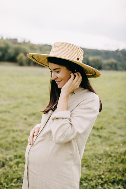 Portrait of a pregnant woman with brunette hair outdoors in nature wearing a straw hat