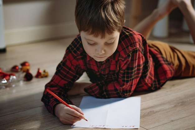 Portrait of preschooler boy in plaid shirt laying on the floor writing christmas letter to santa