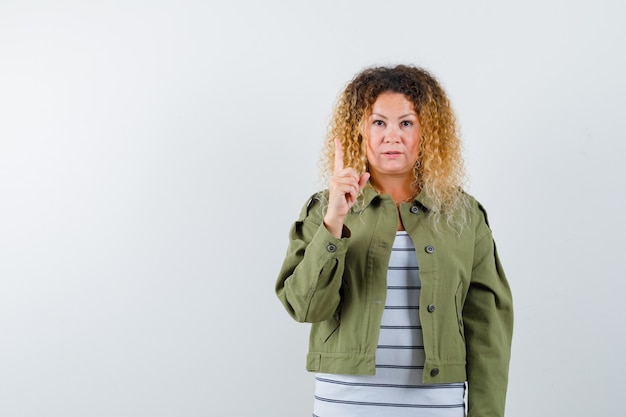 Portrait of pretty blond woman pointing up in green jacket and looking pensive front view