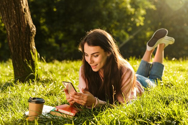 Portrait of pretty brunette woman smiling and using cellphone while lying at grass in green park on sunny day