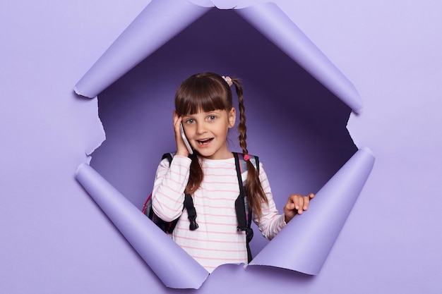 Portrait of pretty Caucasian school girl with braids wearing backpack holding smart phone talking on cell phone looking at camera posing in violet paper hole