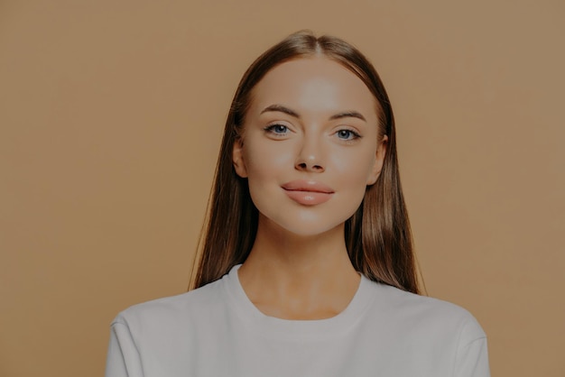 Portrait of pretty European woman with long hair looks confidently ar camera has healthy glowing skin makeup wears casual jumper isolated over brown studio wall Beauty and wellness concept