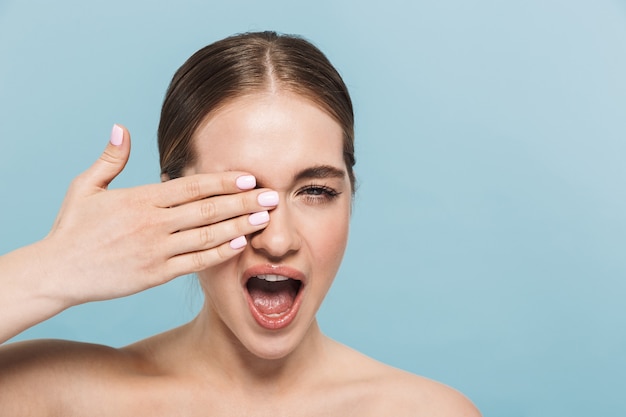 Portrait of a pretty excited young woman posing isolated over blue wall covering eye winking.