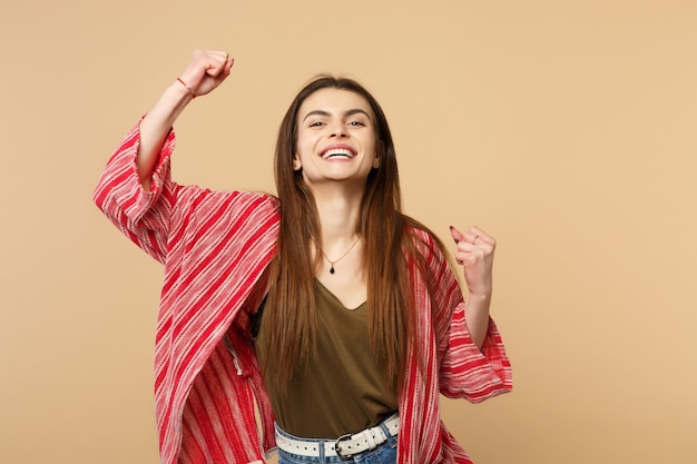 Portrait of pretty joyful young woman in casual clothes clenching fists like winner isolated on pastel beige wall background in studio. People sincere emotions, lifestyle concept. Mock up copy space.