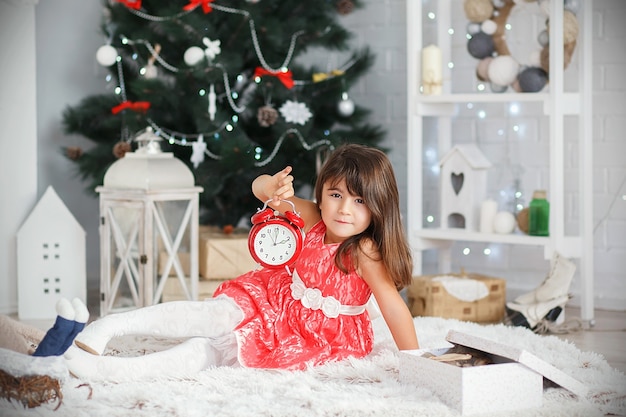 Portrait of a pretty little brunette girl holding a red alarm clock in the hands in the interior with Christmas decorations