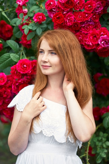 Portrait of a pretty redhead girl dressed in a white light dress on a background of blooming roses. 