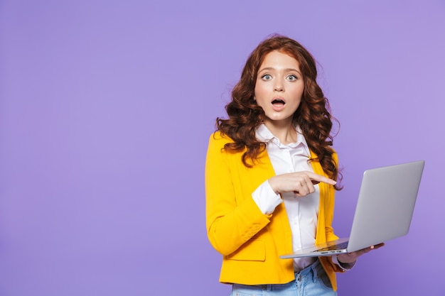 Portrait of a pretty shocked young redheaded woman standing over violet, using laptop